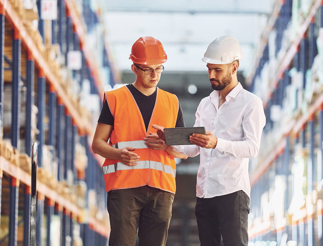 Two men wearing hard hats stand in a warehouse, surrounded by shelves and industrial equipment.